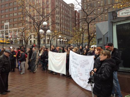 Photo : foule au bord du chapiteau de la Place D'Youville, tenant deux bannières blanches. La foule regarde une pièce de théâtre de rue.*